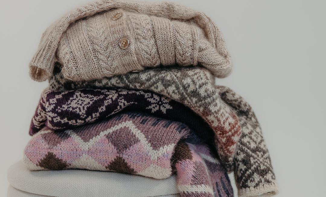 A stack of folded colourwork sweaters in oatmeal, plum, pink and brown tones on a pale stool