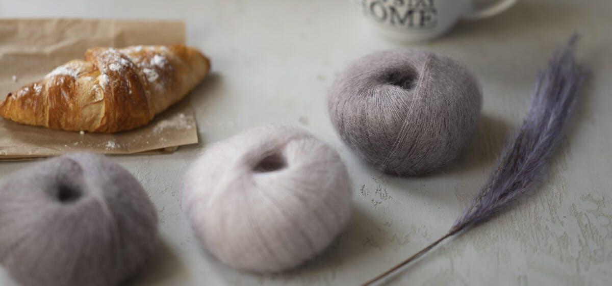 Three balls of fluffy mohair yarn in lilac and dusty pink tones beside a croissant and dried grass
