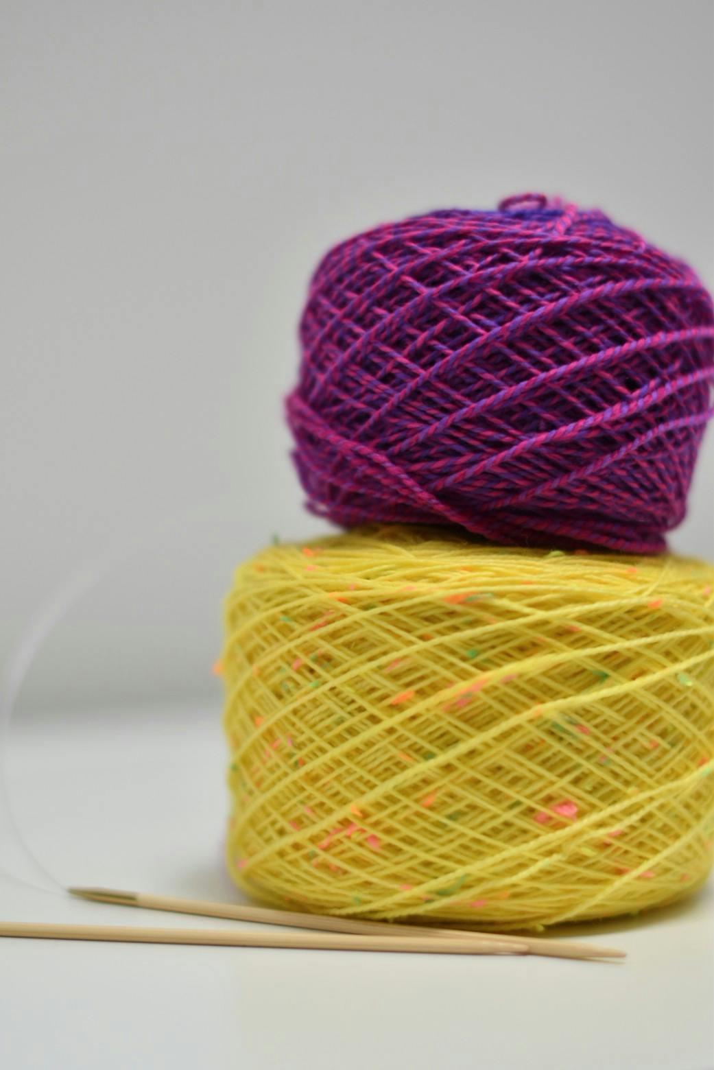 Cake of purple-pink Spincycle yarn stacked on a yellow neon-flecked Moondrake yarn cake with bamboo DPNs alongside